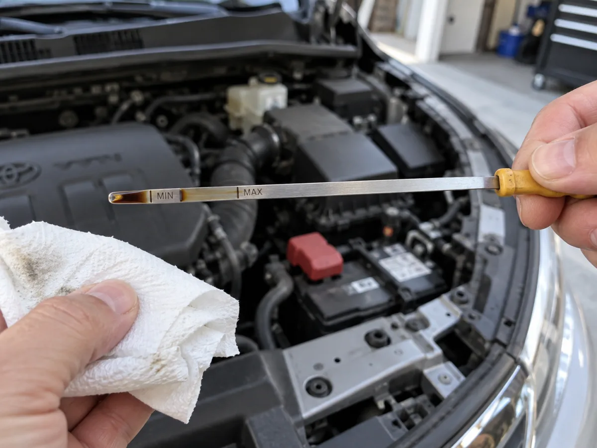 A hand holding an engine oil dipstick between the MIN and MAX marks, with a paper towel used to wipe it clean over an open engine bay.